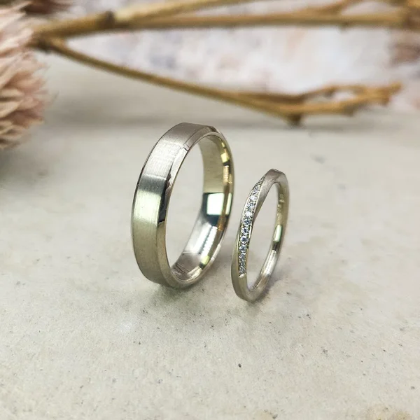 Zwei silberne Eheringe auf hellem Untergrund; einer ist schlicht und breiter, der andere schmaler und mit kleinen Diamanten besetzt. Im Hintergrund sind getrocknete Blumen verschwommen.