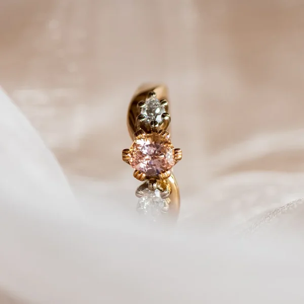 Eine Nahaufnahme eines goldenen Rings mit einem rosafarbenen Edelstein in der Mitte und zwei kleineren klaren Steinen, der auf einem weichen, weißen, durchsichtigen Stoff ruht.